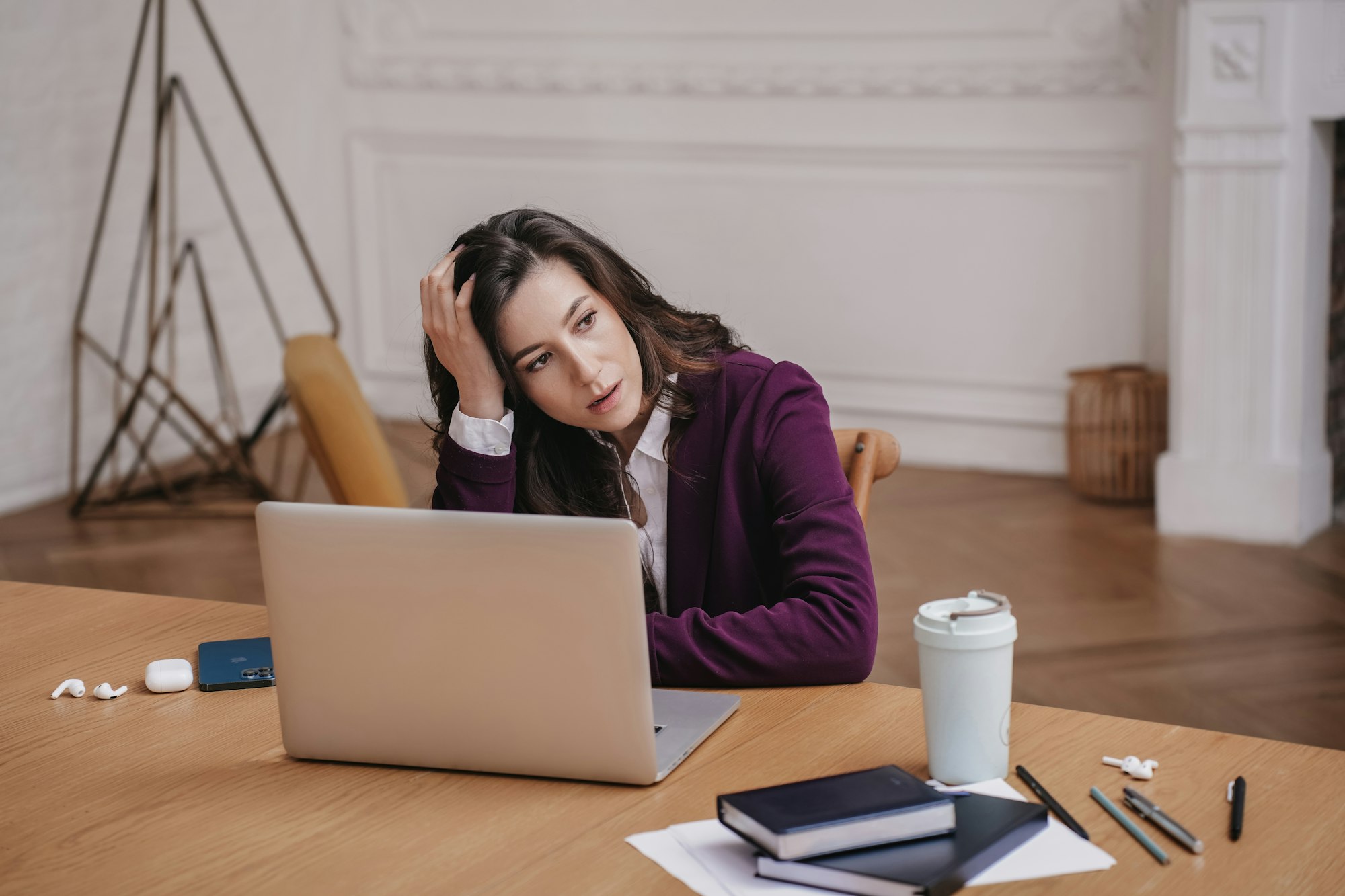 Exhausted hispanic businesswoman in burgundy suit sitting at desk with laptop, notebook, coffee